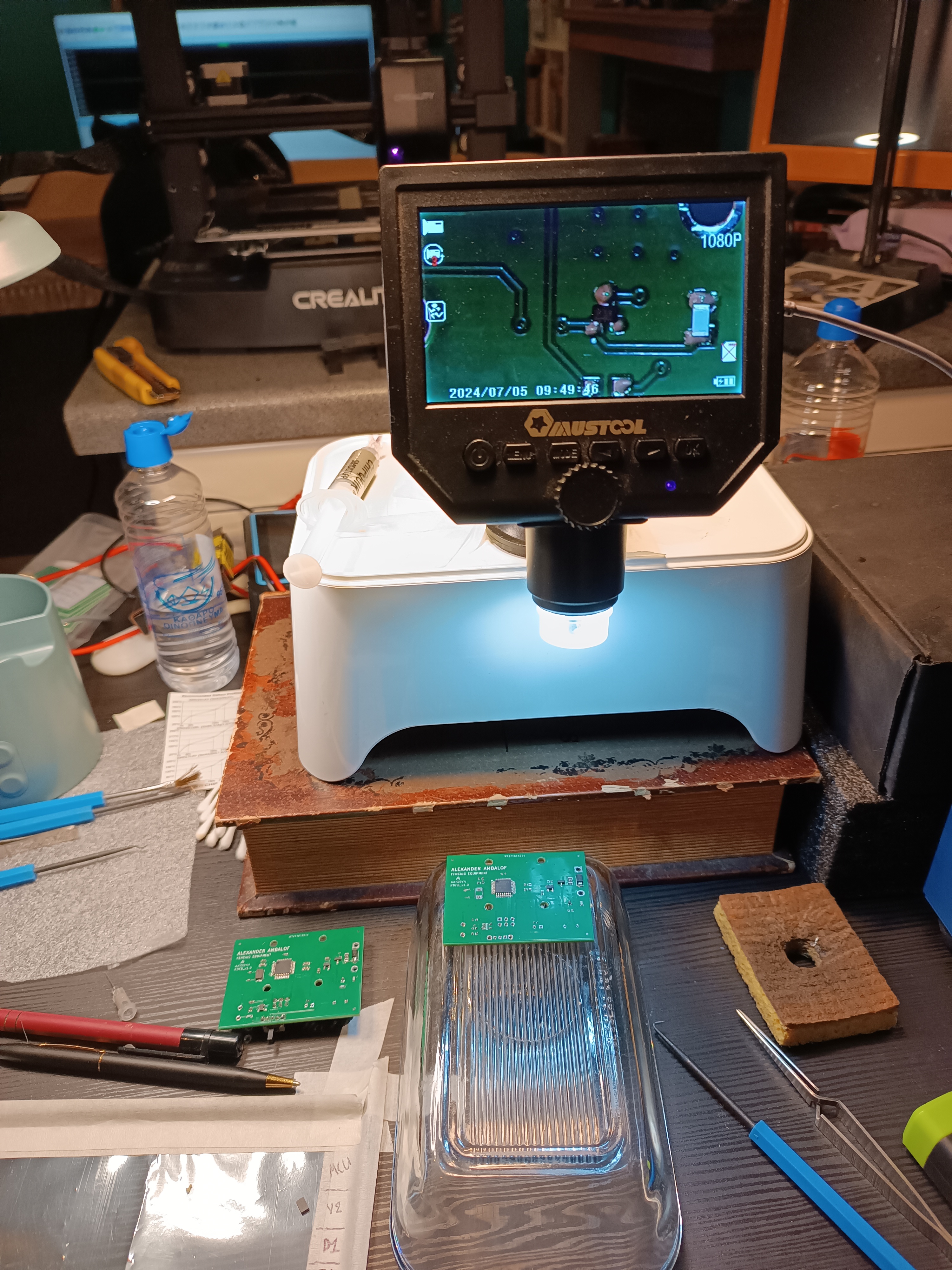 Workbench: digital microscope inspecting populated PCB, two boards in foreground, 3D printer behind