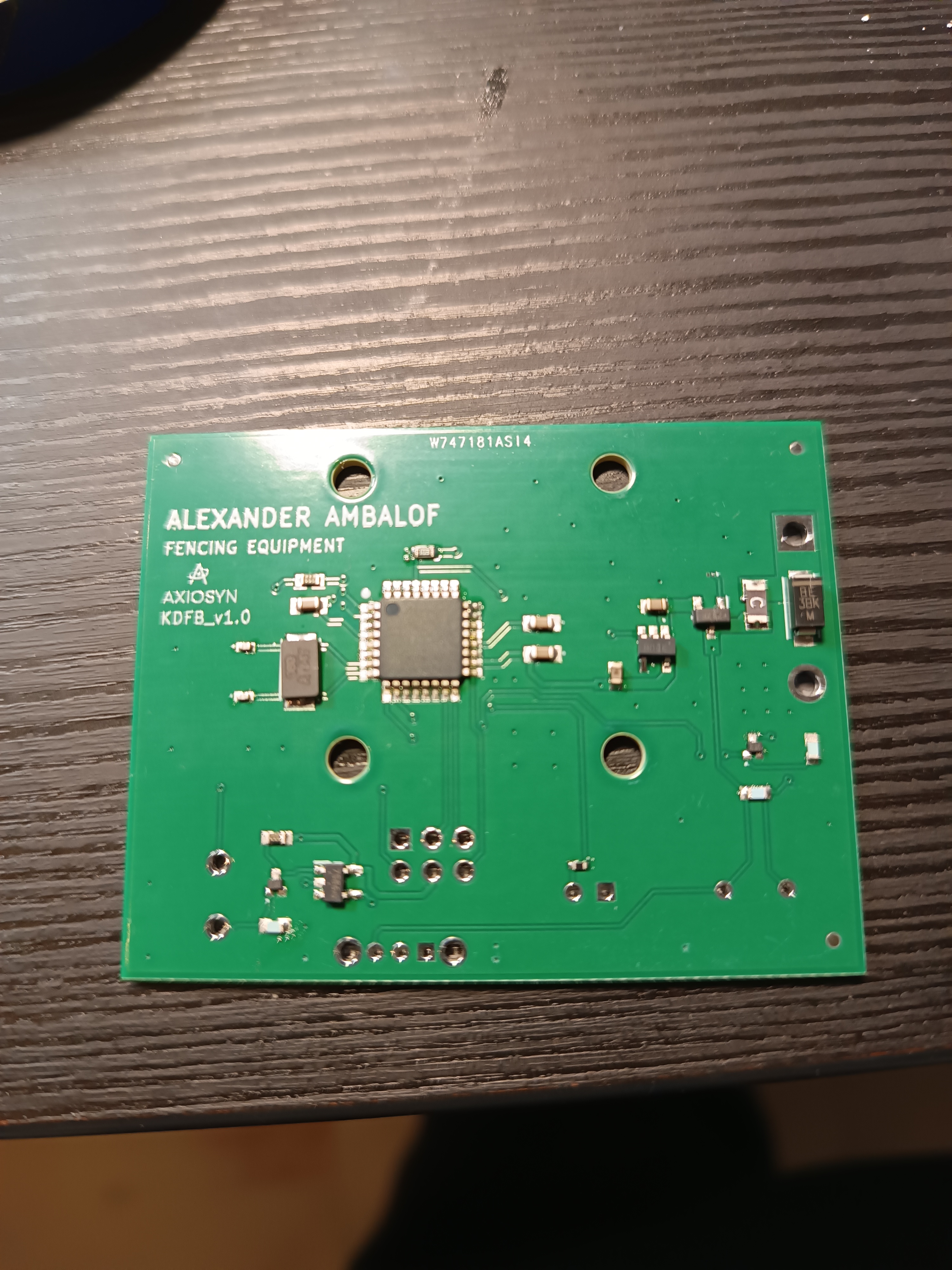 Top-down view of the assembled custom PCB showing the QFN microcontroller, crystal, regulator, TVS diode, and AXIOSYN silkscreen branding